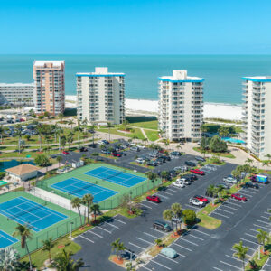 Aerial view of a beachfront resort with tall white condo towers, palm trees, a parking lot, and blue tennis courts by the ocean.